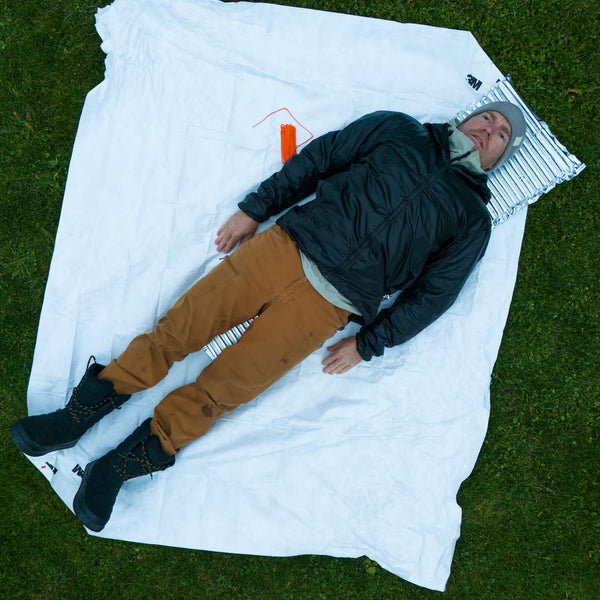 Adult lying on the white Tyvek Bruce Wayne Rescue Tarp on grass, showing the tarp's full size for a patient