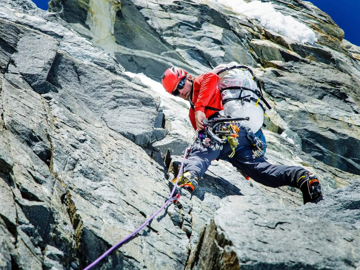 Alpinist in a red helmet leading steep alpine rock, rope trailing below on a sunlit face