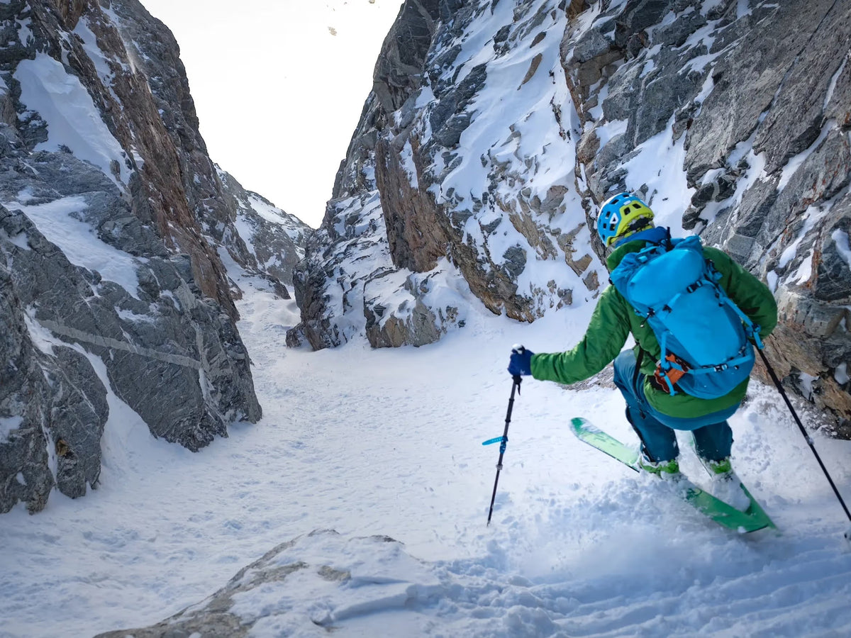 Backcountry skier making a turn in a narrow rock walled couloir with a blue touring pack