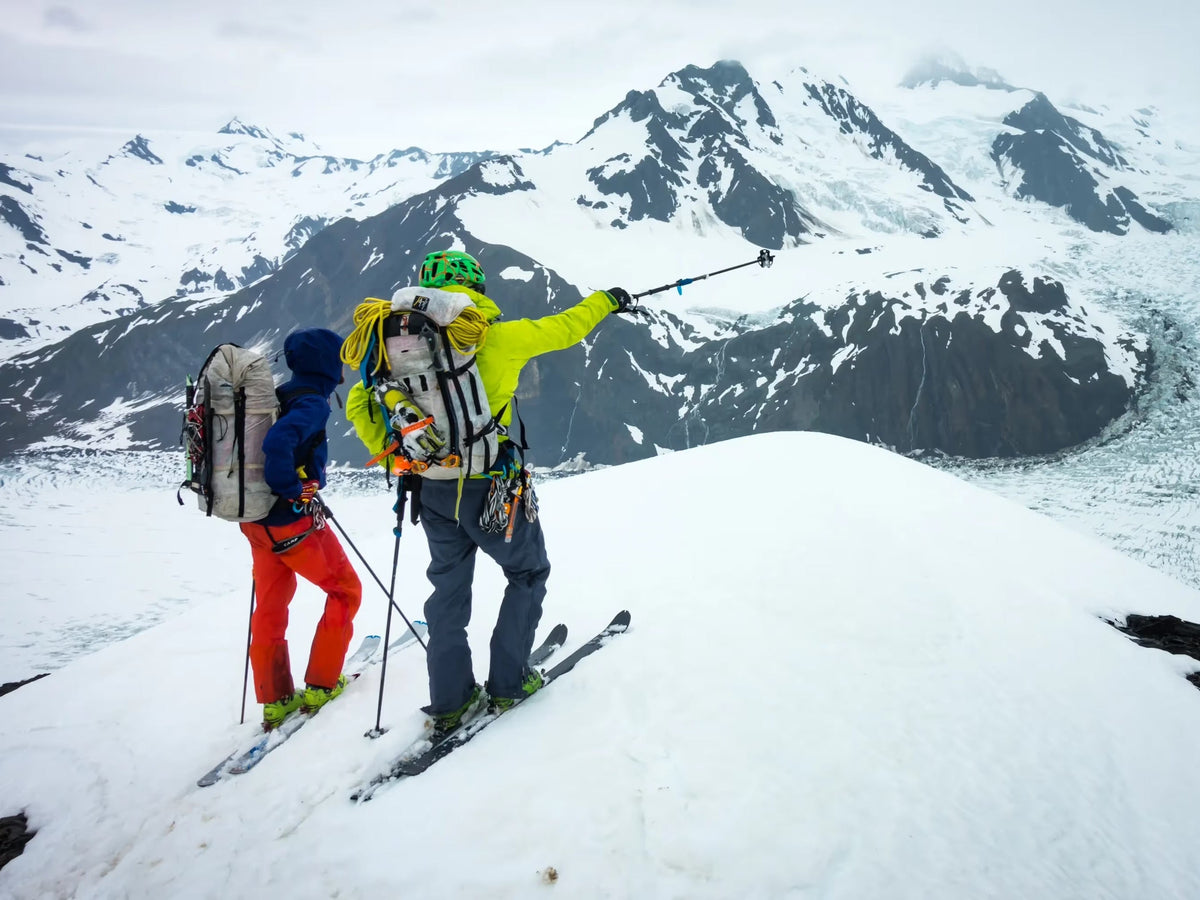 Two ski tourers on a summit ridge, one pointing across a glaciated range toward the next objective