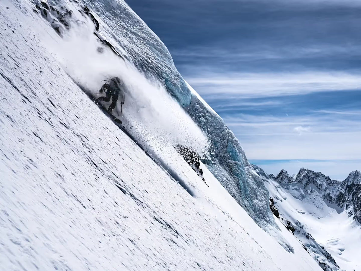 Splitboarder riding a steep snow face throwing spindrift, jagged peaks along the horizon