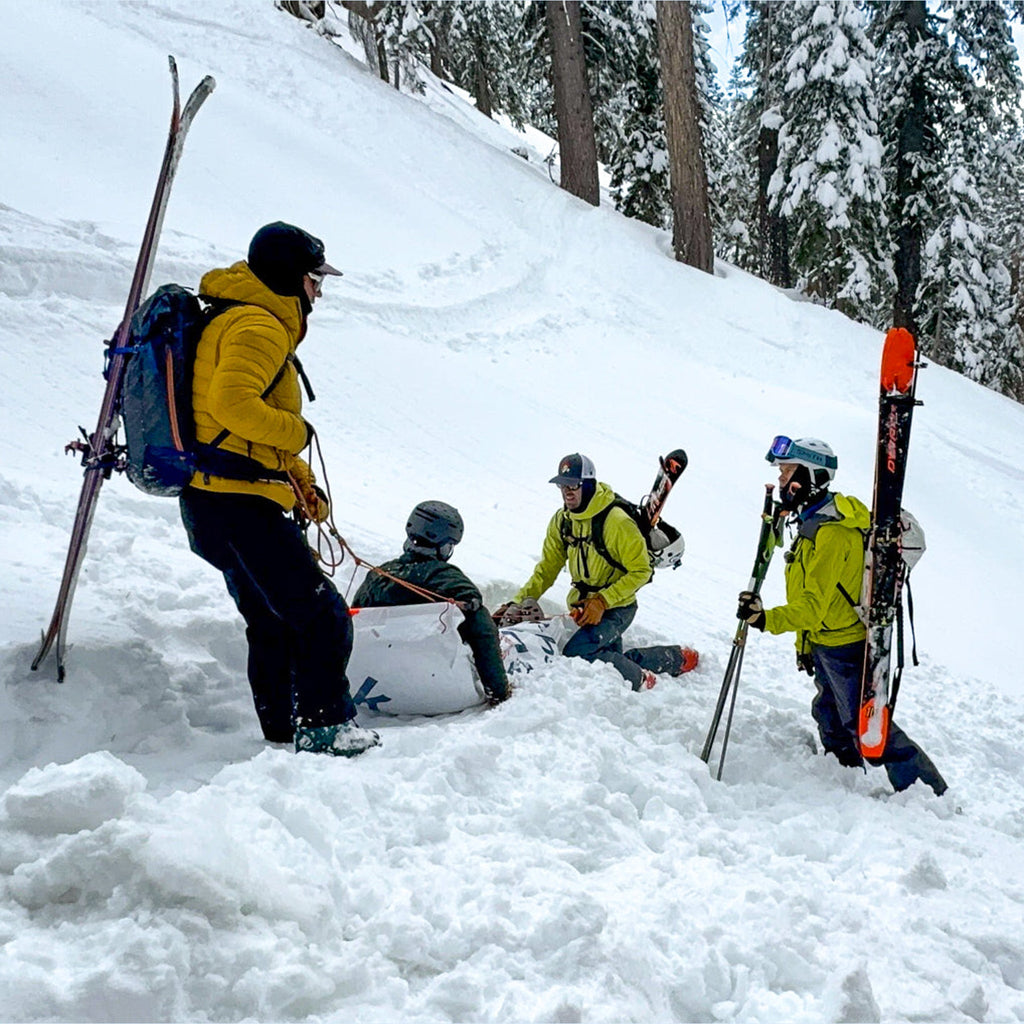 Real backcountry rescue in progress: three skiers assist an injured partner sitting in the snow below snowy trees